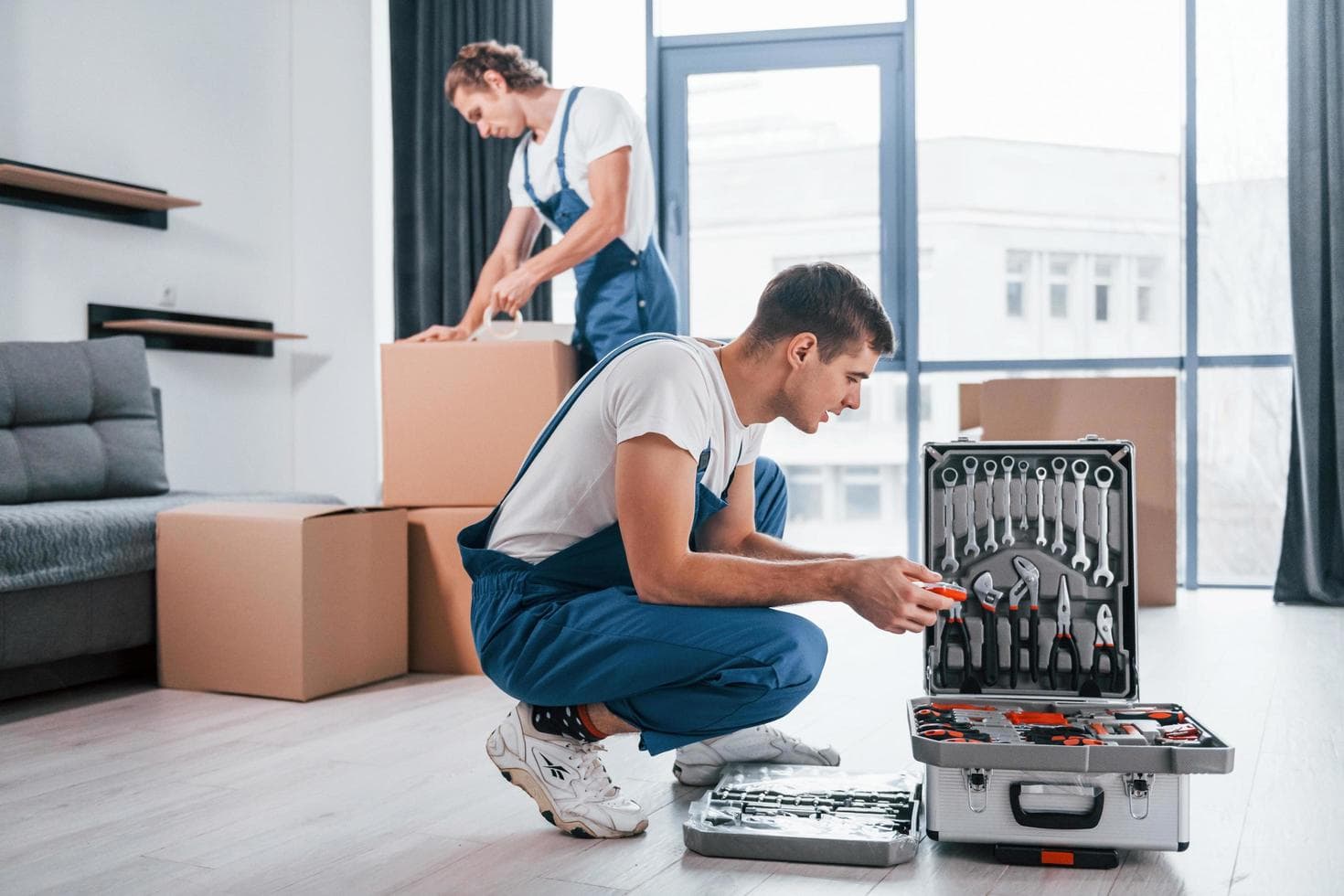 Two young movers in blue uniform using tools from a case while working indoors in a room